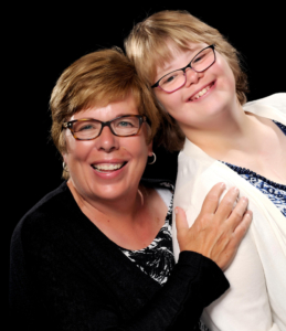 A loving mother and daughter smile and pose for a picture. They both have blonde hair that is cut short in pixie cuts, and they are wearing glasses with dark rectangular frames. The mother is wearing a black cardigan and earrings, while the daughter is wearing a white cardigan over a blue floral blouse. The daughter has Down syndrome.